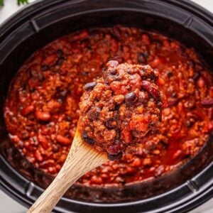 A wooden spoon holds a scoop of slow cooker chili above a slow cooker, with beans and meat simmered in a rich tomato sauce.