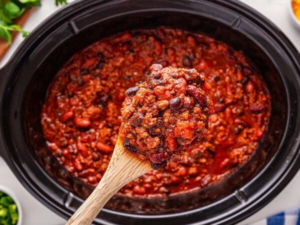 A wooden spoon holds a scoop of slow cooker chili above a slow cooker, with beans and meat simmered in a rich tomato sauce.