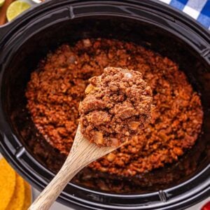 A wooden spoon holds seasoned Slow Cooker Taco Meat over a slow cooker, surrounded by taco shells and fresh toppings.