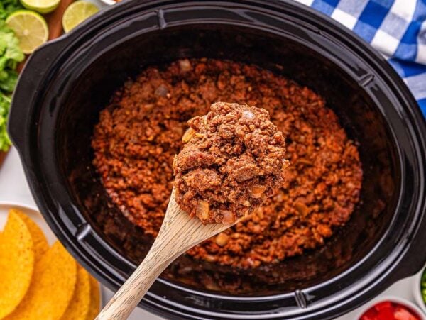 A wooden spoon holds seasoned Slow Cooker Taco Meat over a slow cooker, surrounded by taco shells and fresh toppings.
