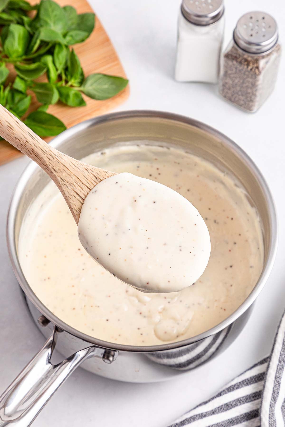 A wooden spoon holds creamy white sauce over a saucepan, with basil and salt and pepper shakers nearby.