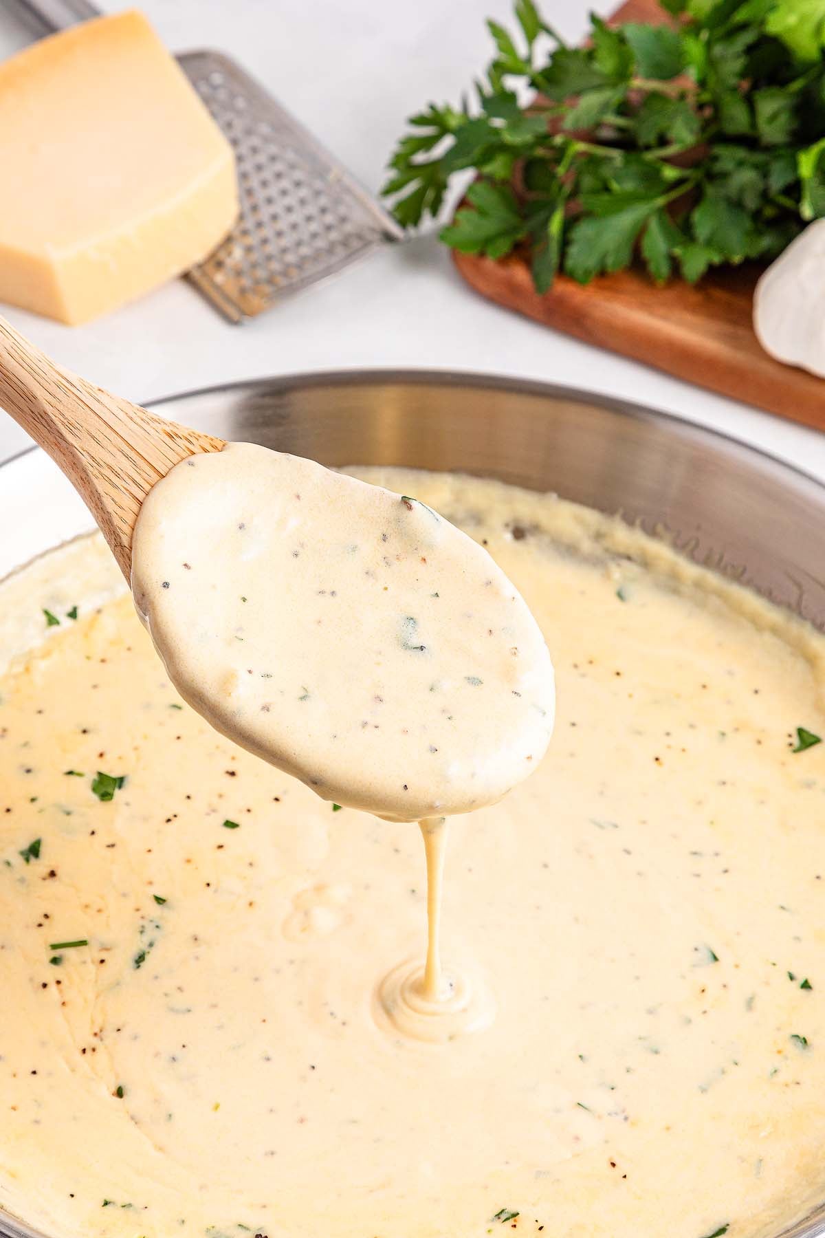 Wooden spoon holds creamy Alfredo sauce above a pan, with herbs, cheese, and parsley in the background.