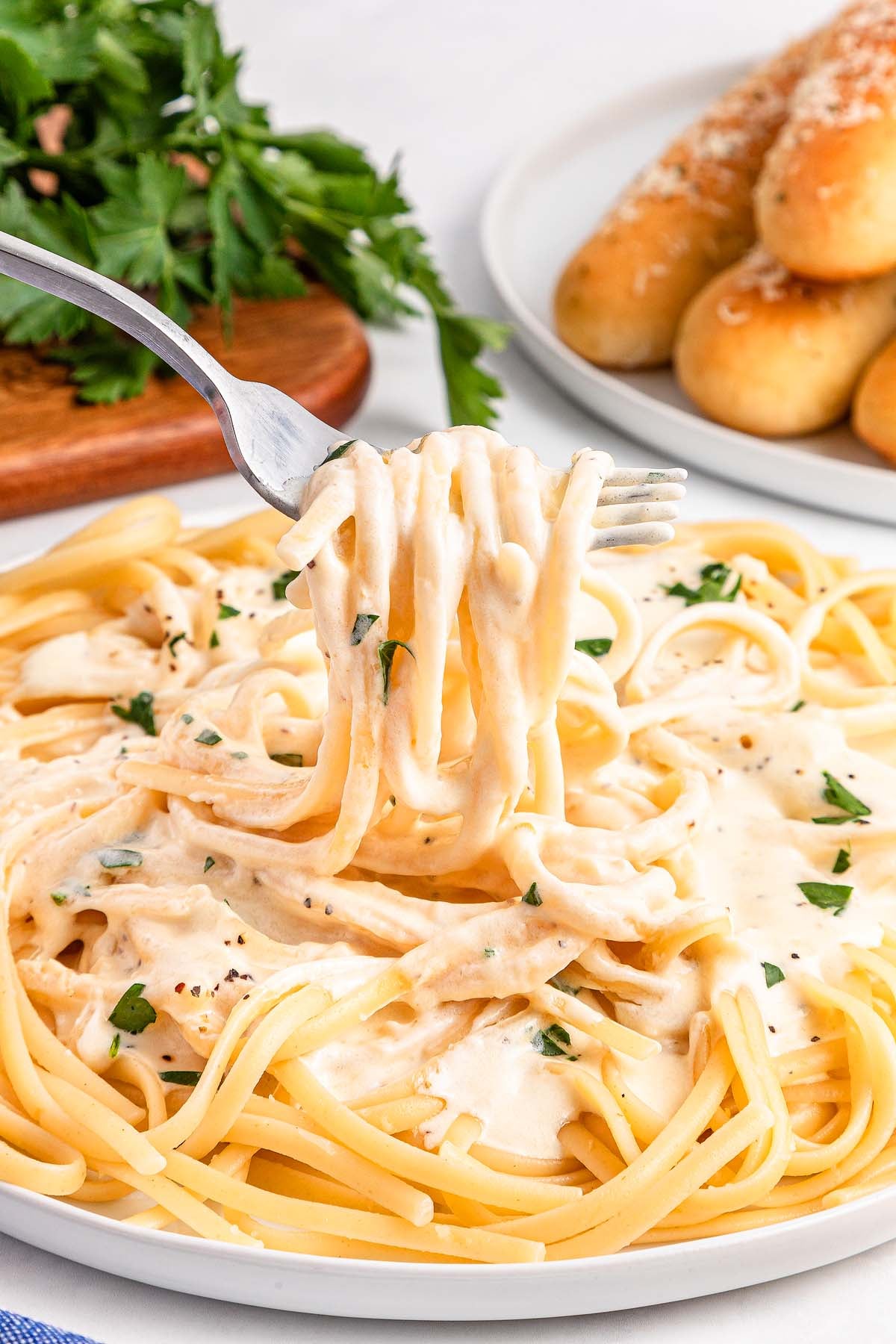 A fork lifting creamy Alfredo pasta from a plate, with breadsticks and parsley in the background.