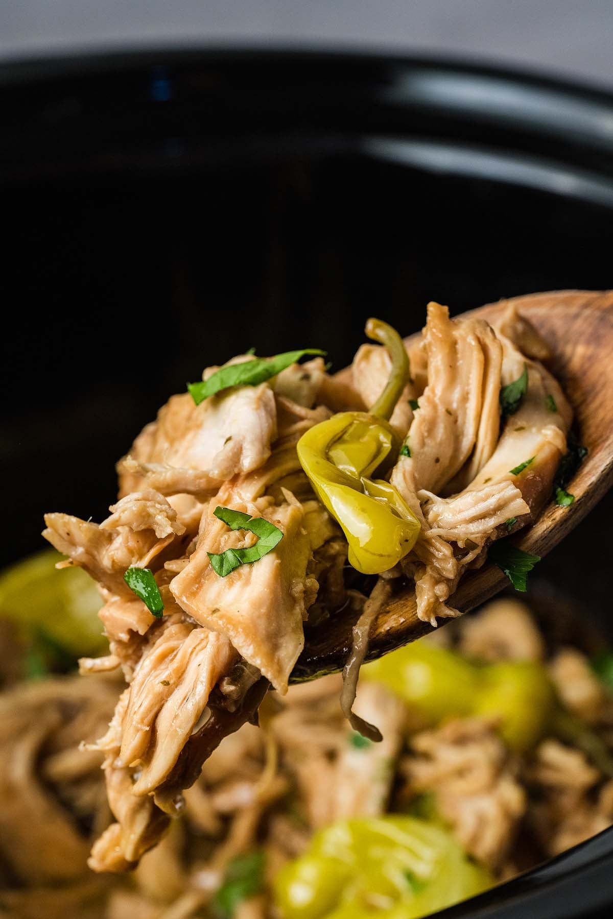 Shredded chicken and pepperoncini on a wooden spoon over a crockpot, garnished with chopped herbs.
