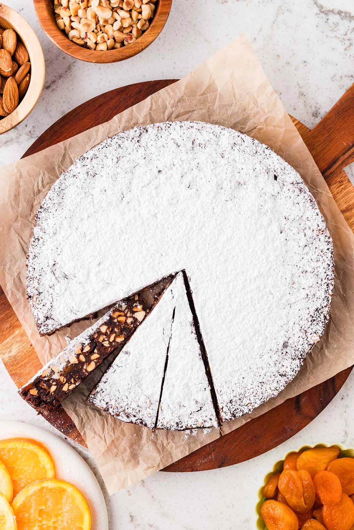 Round chocolate cake with powdered sugar on top, two slices cut, nuts and dried fruit nearby on the table.
