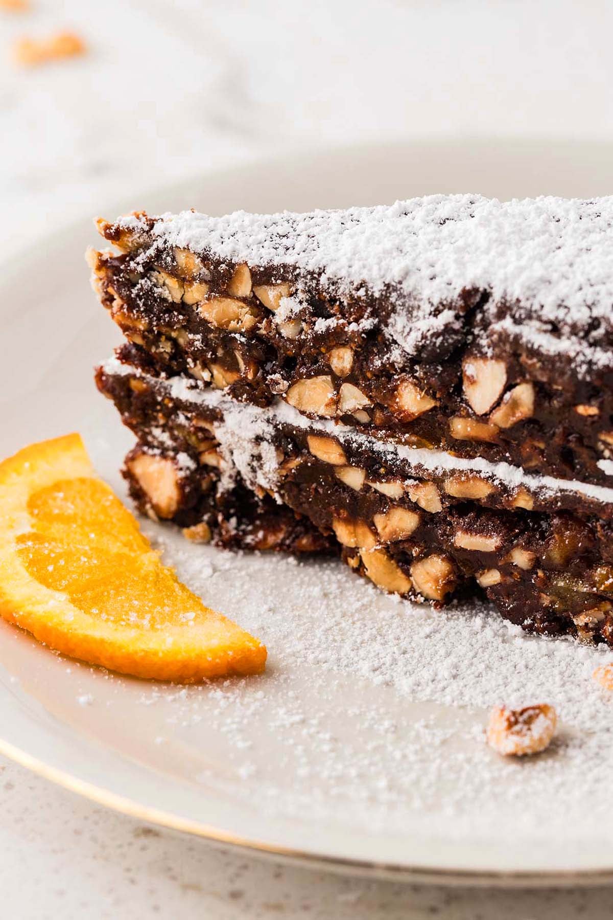 Slice of nutty chocolate cake dusted with powdered sugar, served with an orange slice on a white plate.