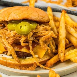 Cowboy burgers come to life with this cheeseburger, piled high with fried onions and jalapeños, served alongside a generous helping of fries on a plate.