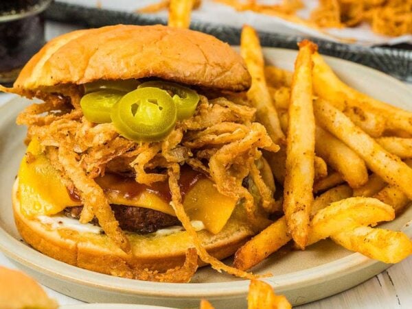 Cowboy burgers come to life with this cheeseburger, piled high with fried onions and jalapeños, served alongside a generous helping of fries on a plate.