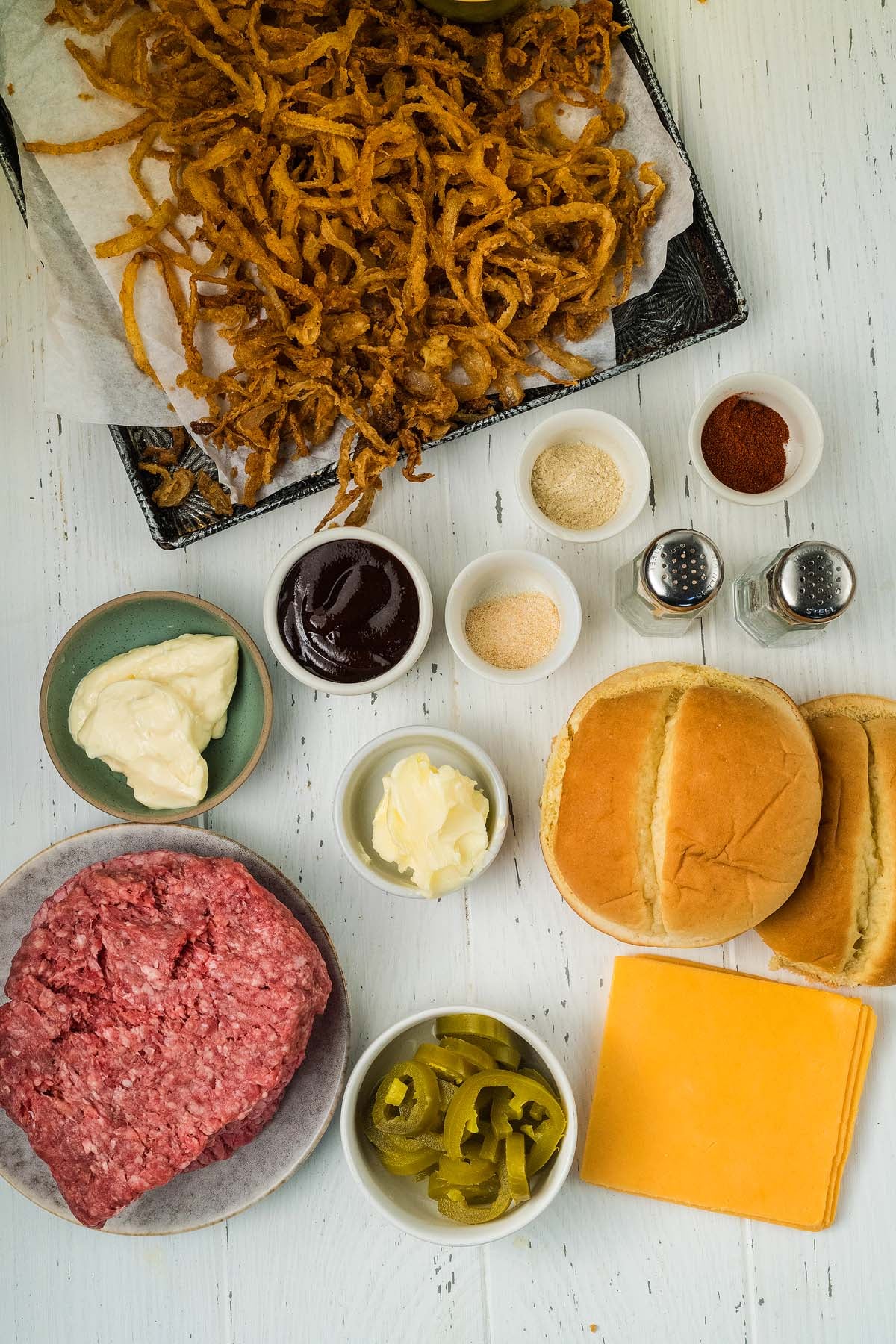 Cowboy burgers ingredients: ground beef, buns, cheese, jalapeños, sauces, spices, mayo, and onions arranged on a white table.