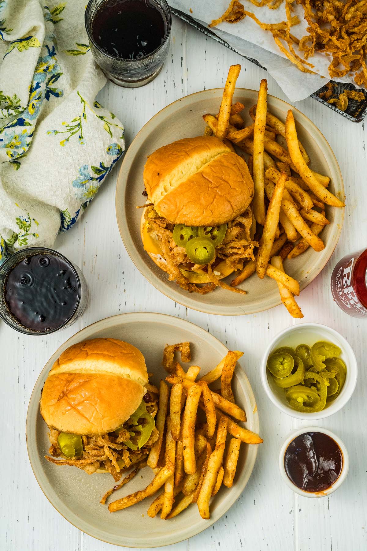 Two plates with cowboy burgers, seasoned fries, jalapeños, barbecue sauce, and two glasses of dark soda.