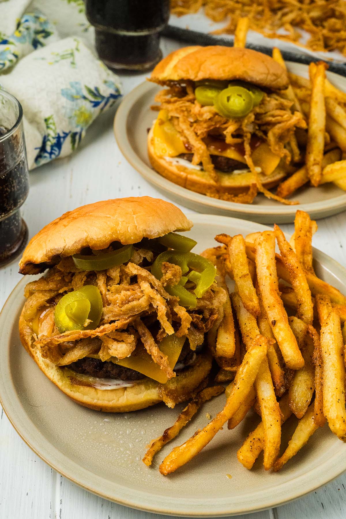 Savor our cowboy burger, topped with crispy fried onions, spicy jalapeños, melted cheese, and a heap of fries on the side—all served up on a plate with a refreshing drink nearby.