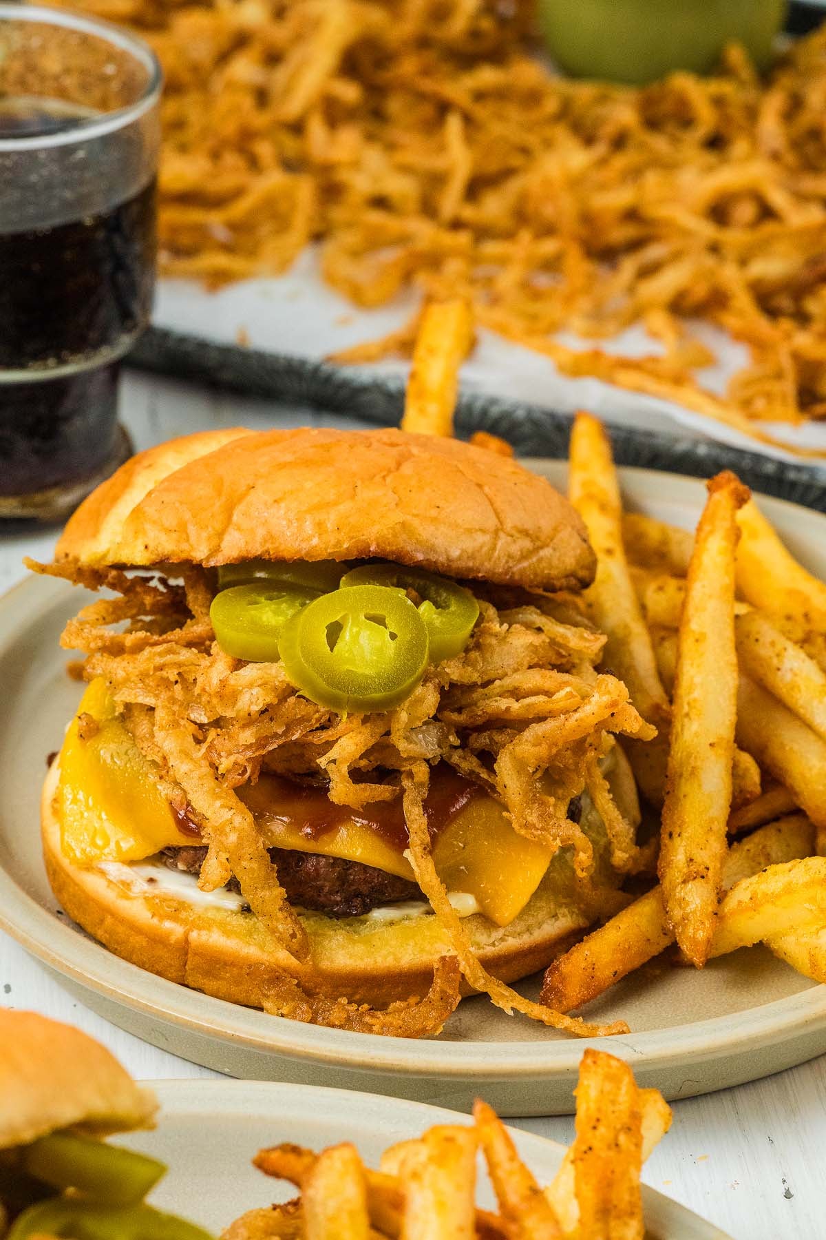Saddle up for flavor with our cowboy burgers—juicy cheeseburger topped with fried onions, jalapeños, served alongside crispy fries and a refreshing drink on a plate.