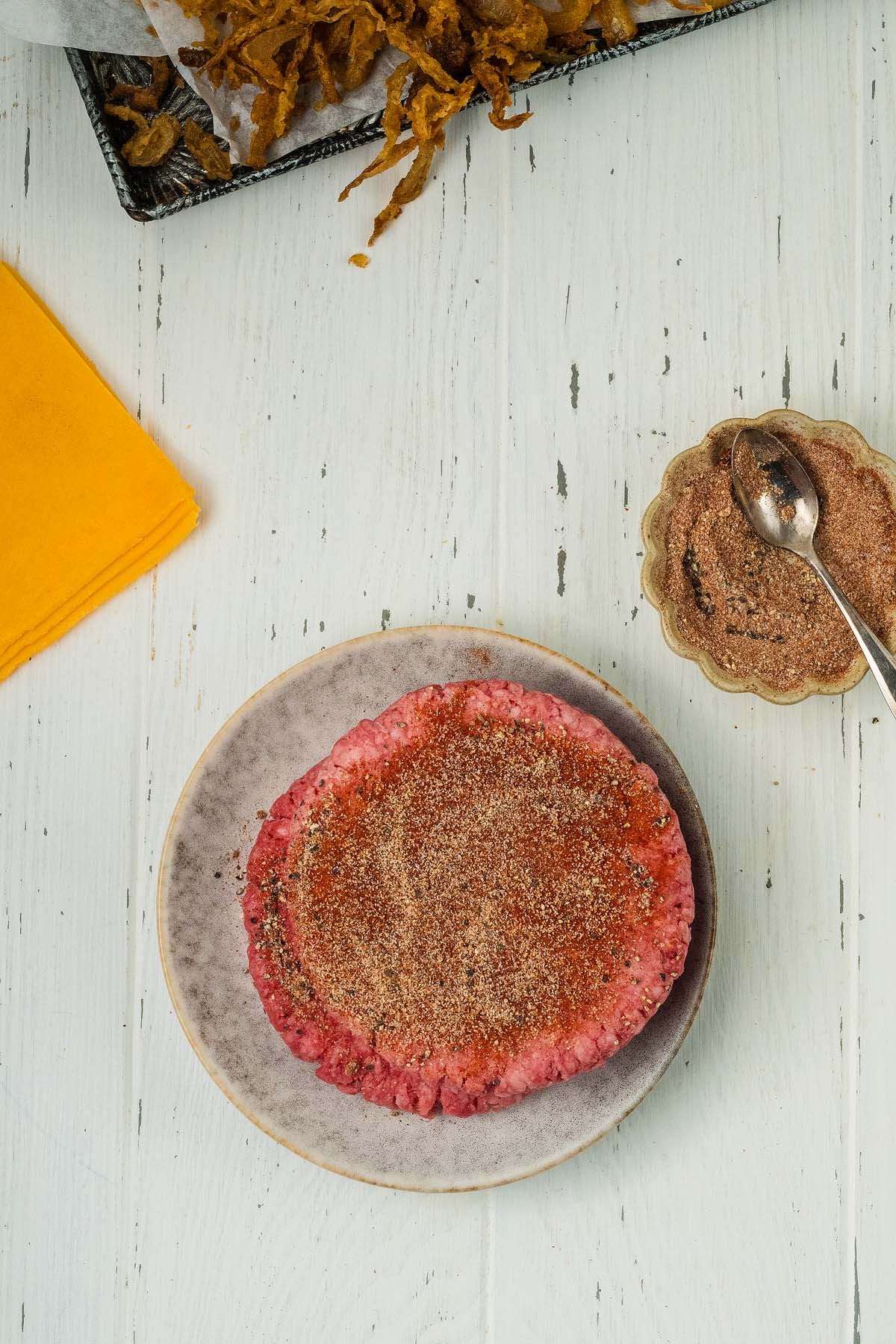 Hamburger meat coated with breadcrumbs on a plate, beside a bowl of spice mix and a yellow napkin.