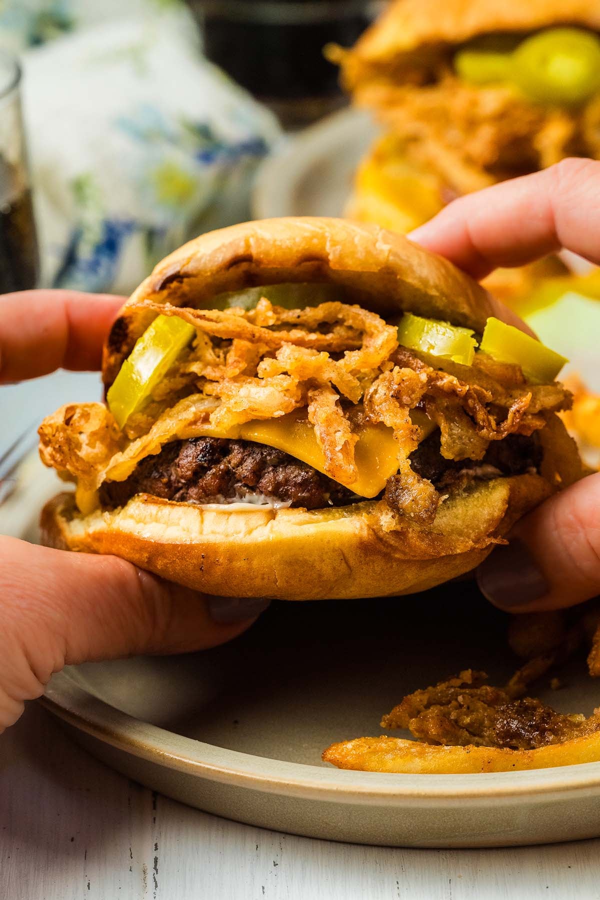 A close-up of hands holding a cowboy burger piled high with fried onions, pickles, and a toasted bun on a plate.