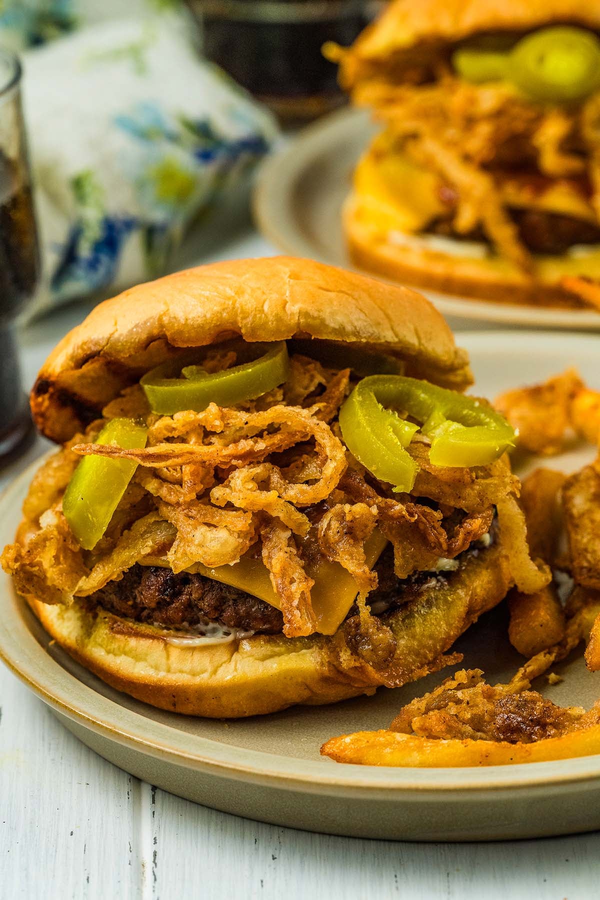 Enjoy one of our signature cowboy burgers, featuring a juicy patty topped with fried onions, pickles, cheese, and served with a side of fries on a beige plate.