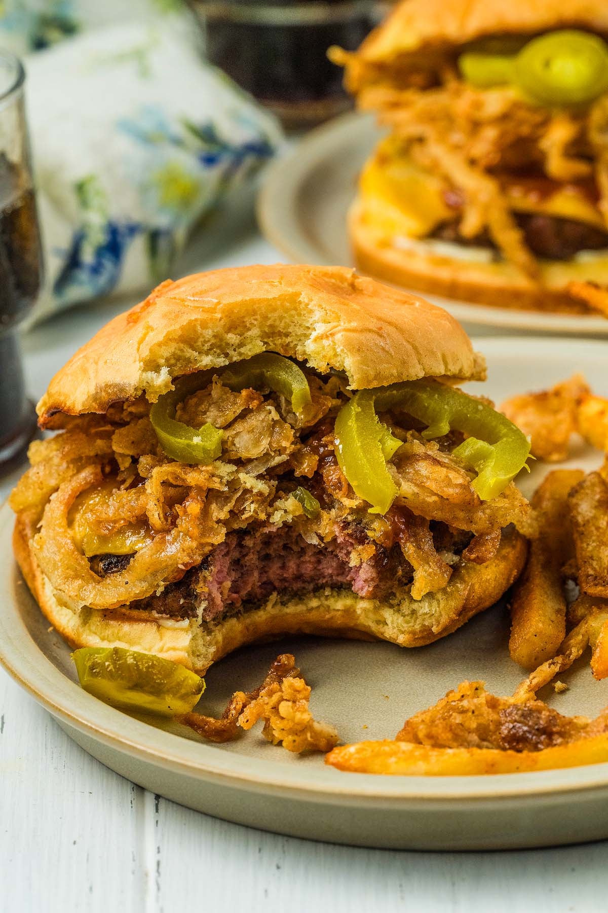 Cowboy burgers with a bite taken out, topped with fried onions and jalapeños, served with fries on a plate.