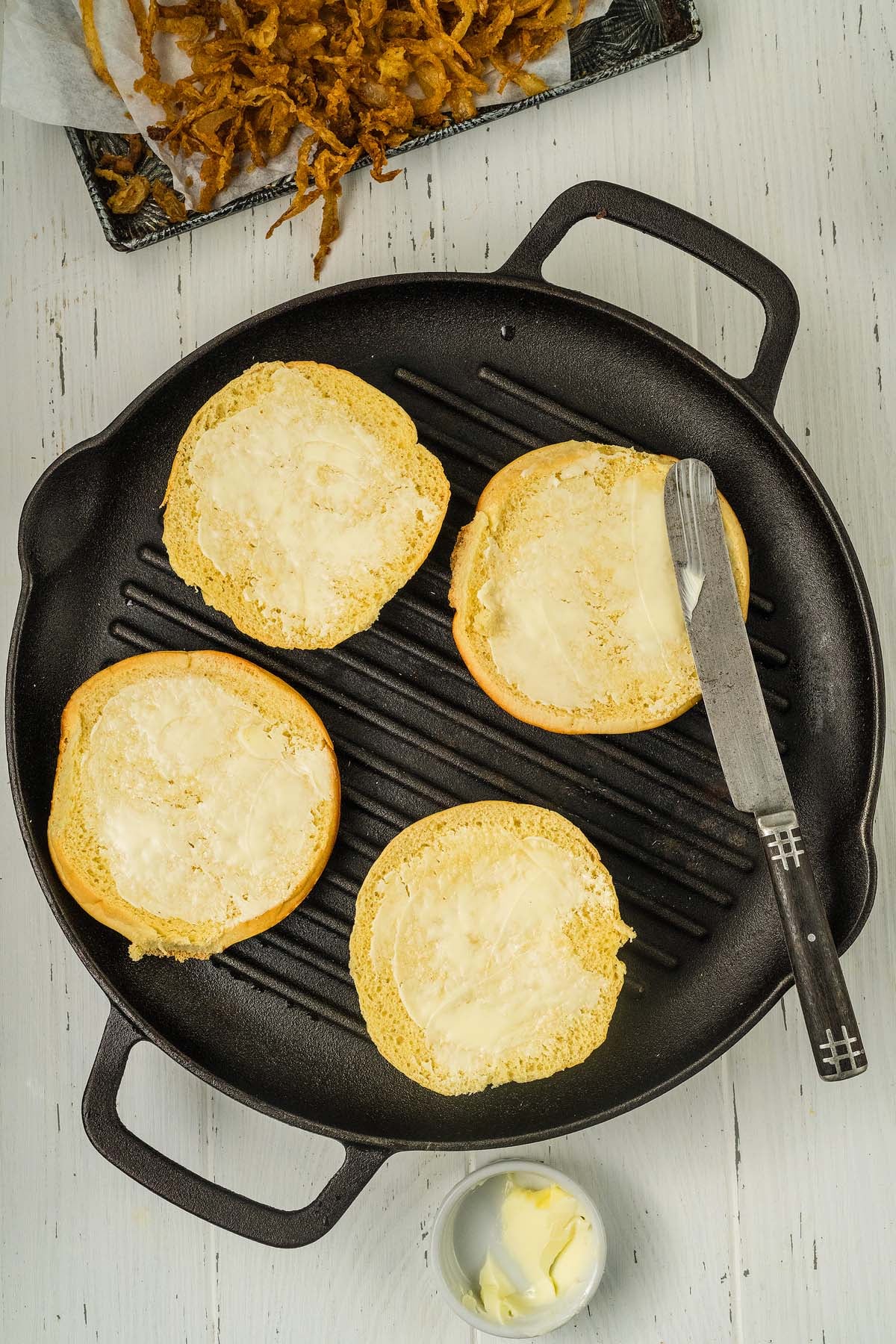 Four buttered burger buns in a grill pan with a butter knife and a small dish of butter nearby, ready to be piled high with juicy cowboy burgers.