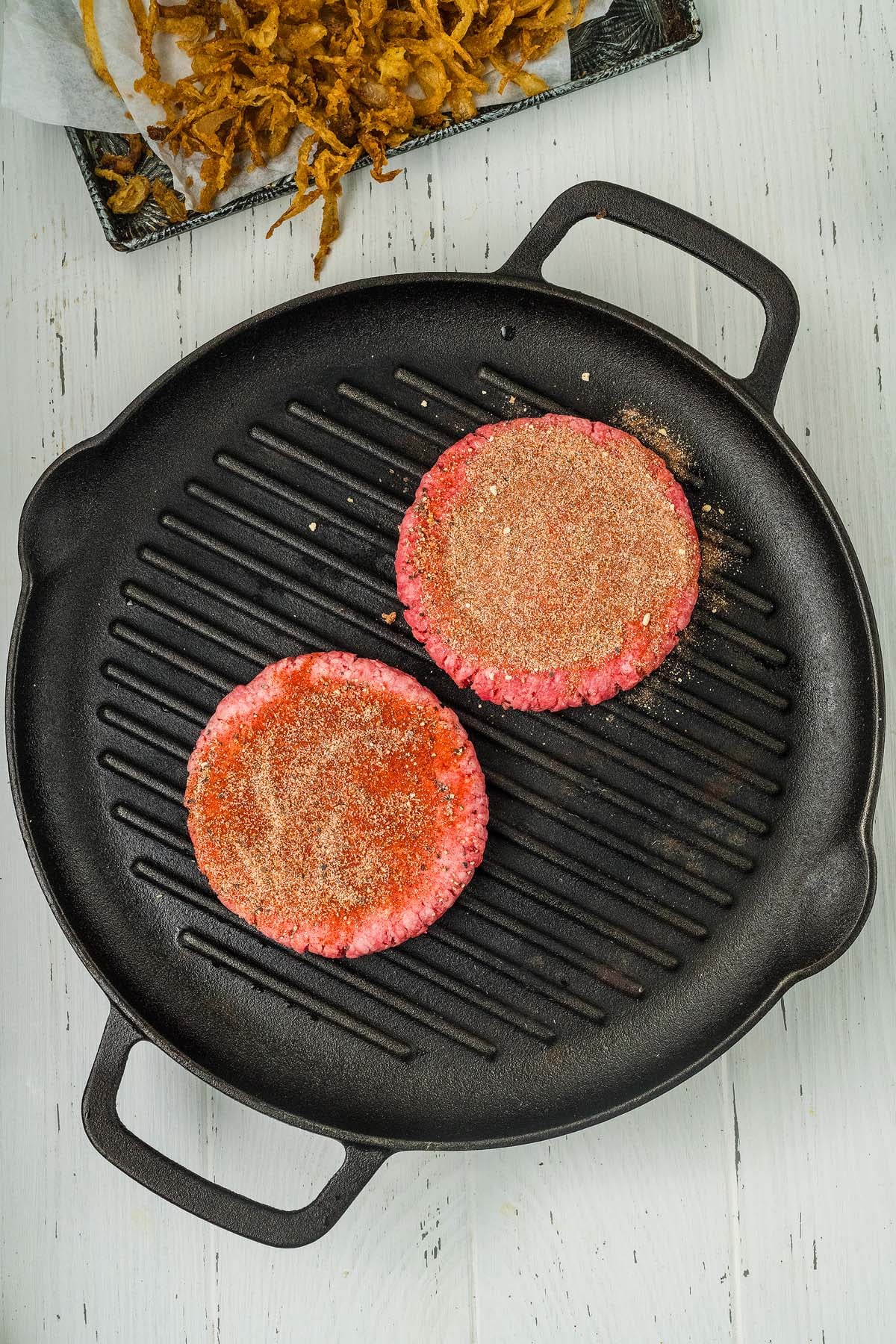 Two raw, seasoned cowboy burger patties on a black grill pan, ready to be cooked, on a light wooden surface.
