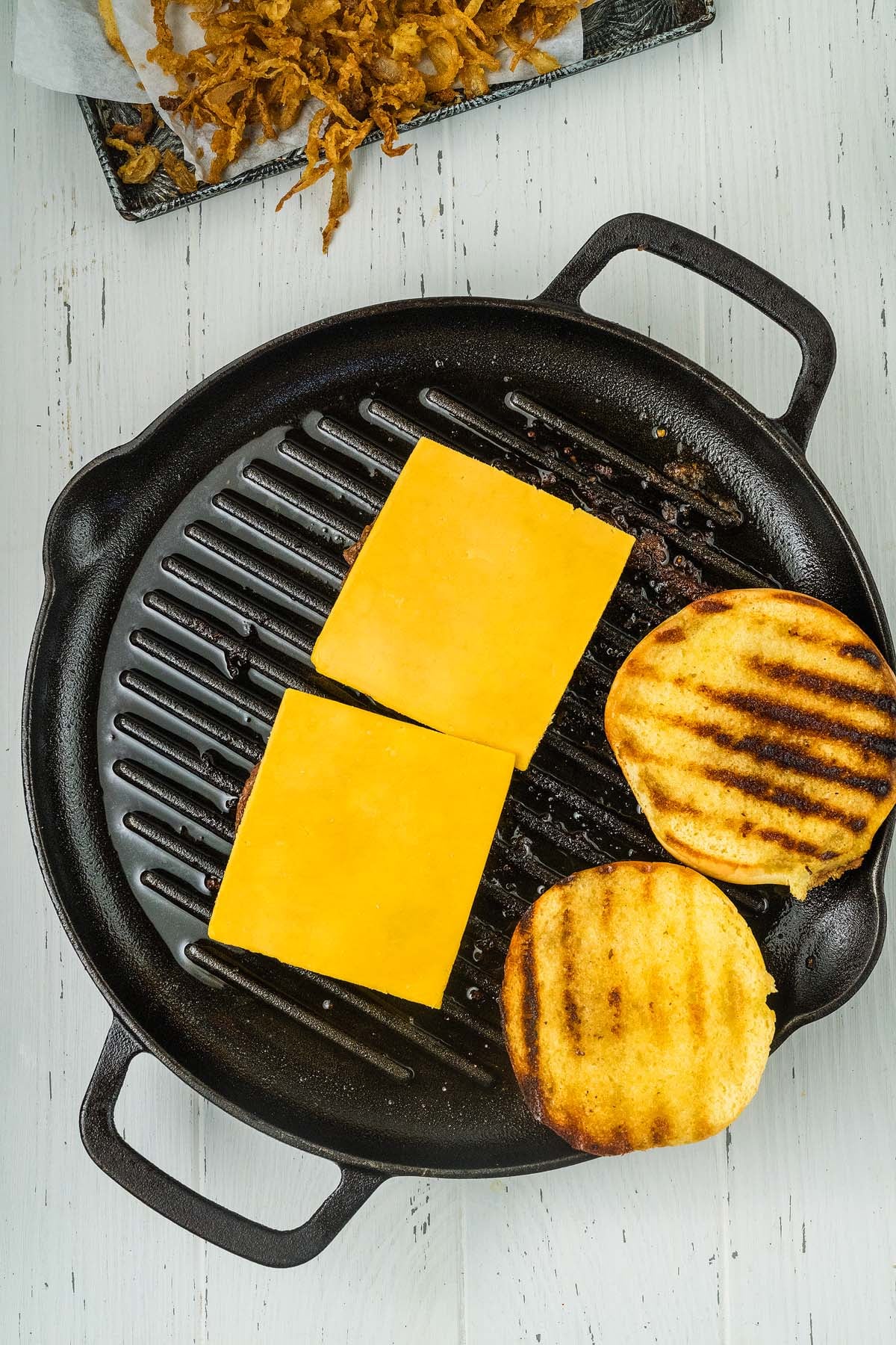 Two cowboy burgers featuring juicy burger patties with melted cheddar cheese and two grilled buns on a black grill pan, overhead view.