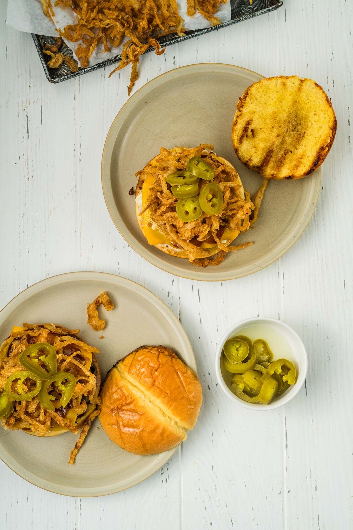Two cowboy burgers with fried onions and jalapeños on plates, with extra fried onions and jalapeños on the side.