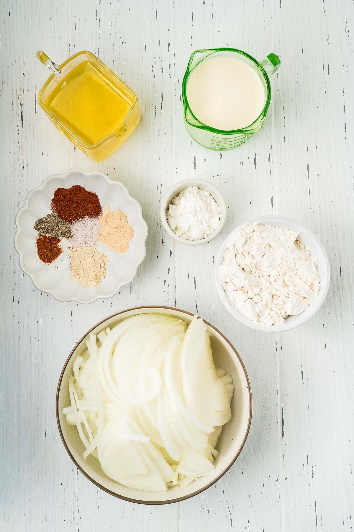 Sliced onions, flour, spices, broth, buttermilk, and cornstarch are arranged in bowls on a white wooden surface&mdash;perfect ingredients for making crispy onion strings.