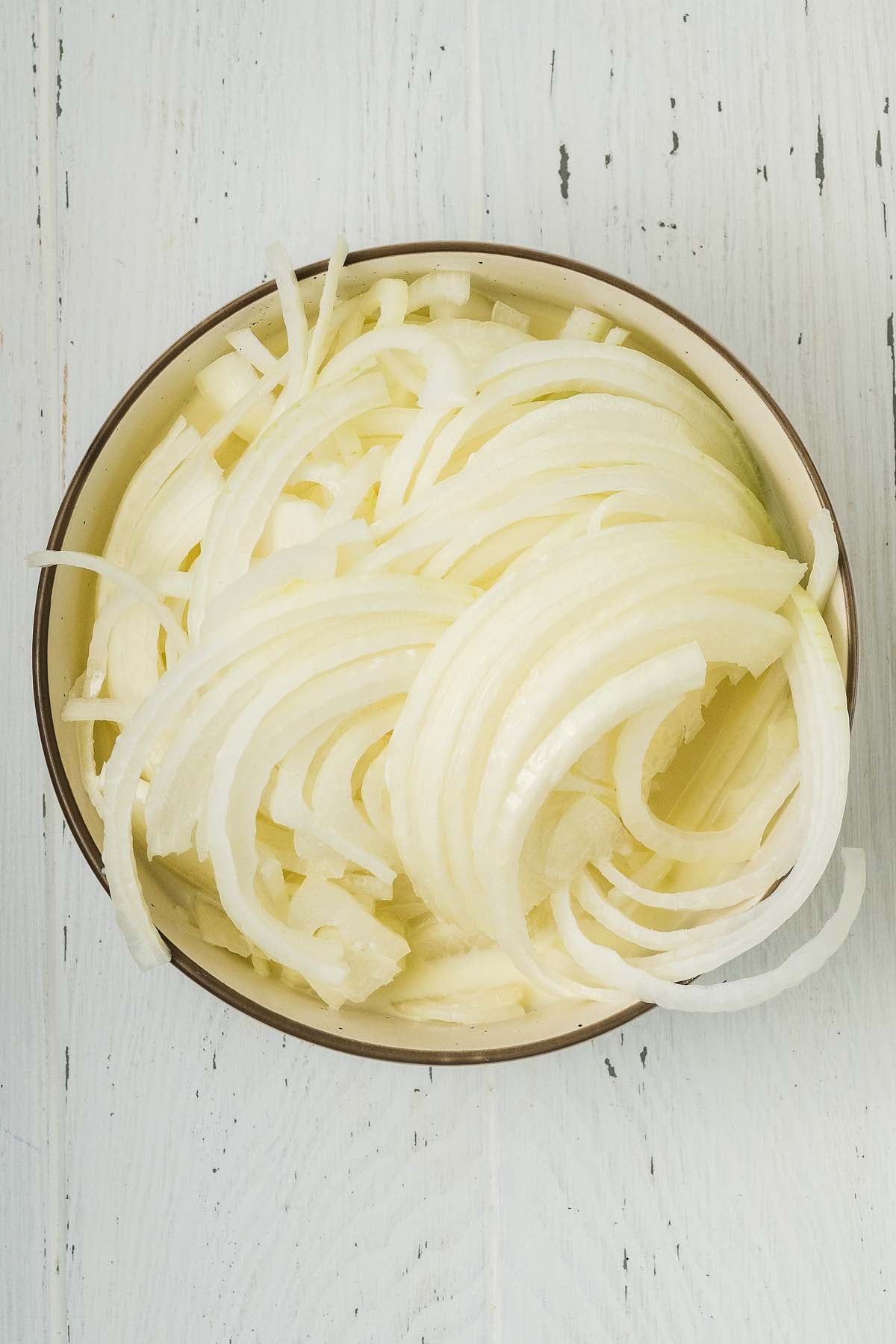 A bowl filled with thinly sliced white onions, perfect for making crispy onion strings, sits on a white wooden surface.