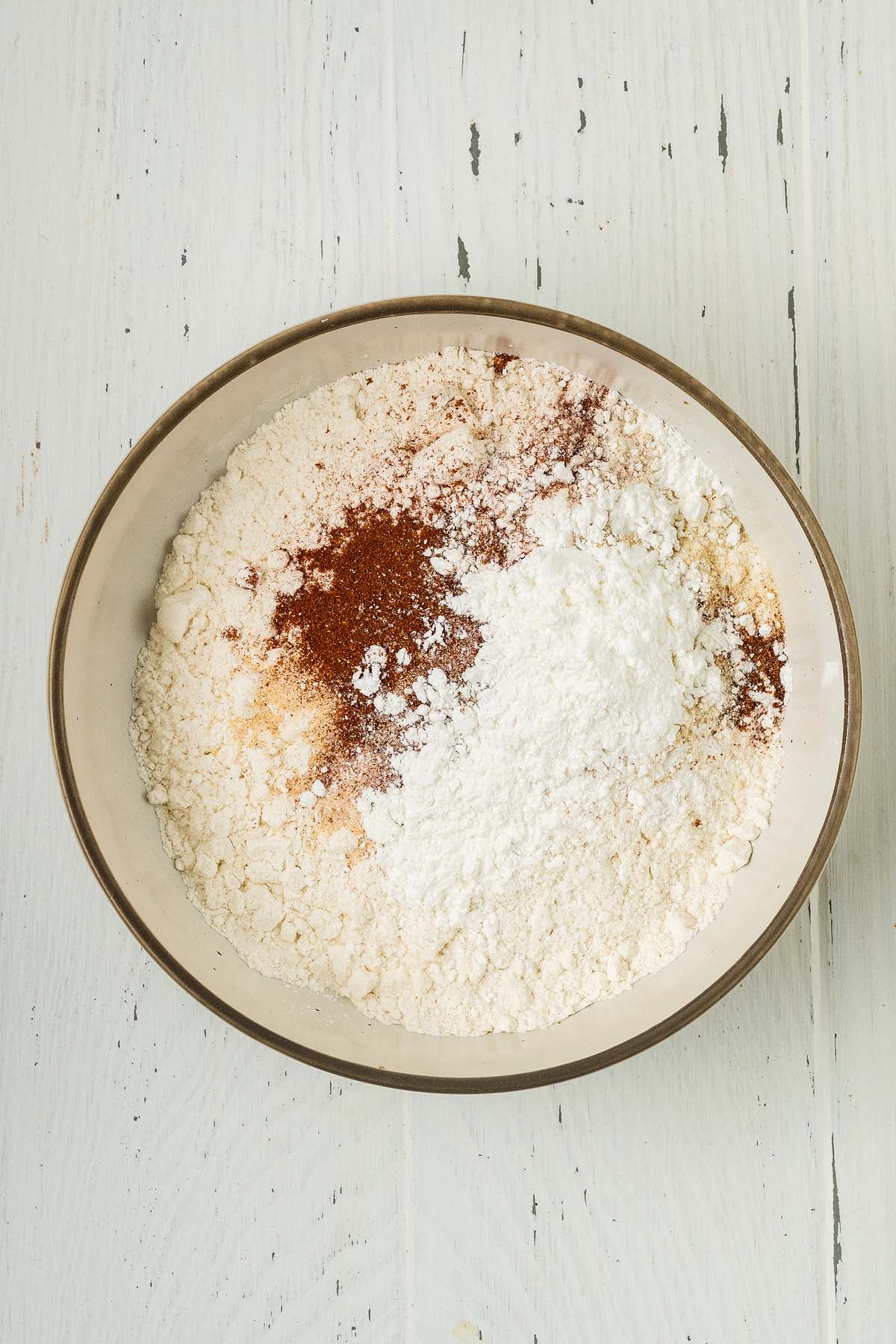 A bowl with dry ingredients and onion strings on a white wooden surface.