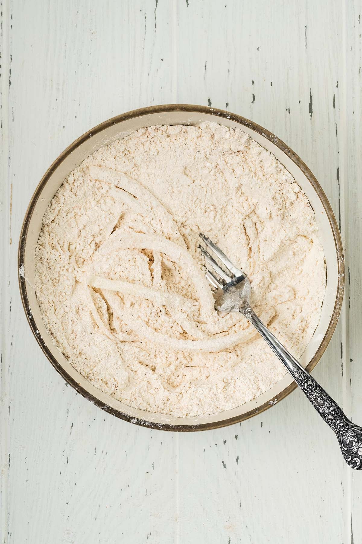 A bowl of flour mixture for crispy onion strings with a fork resting on top, on a white wooden surface.