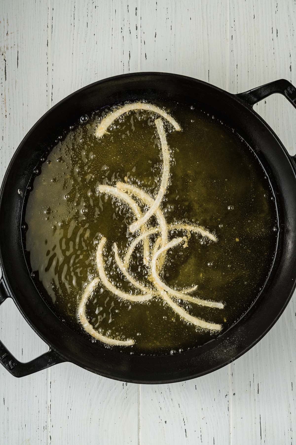 Strips of crispy onion strings frying in hot oil in a black pan on a white wooden surface.