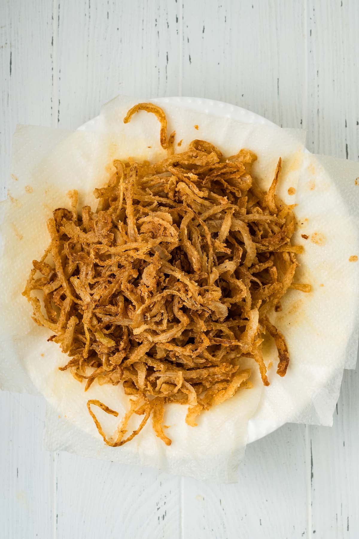 A pile of crispy onion strings rests on a paper towel over a white wooden surface.