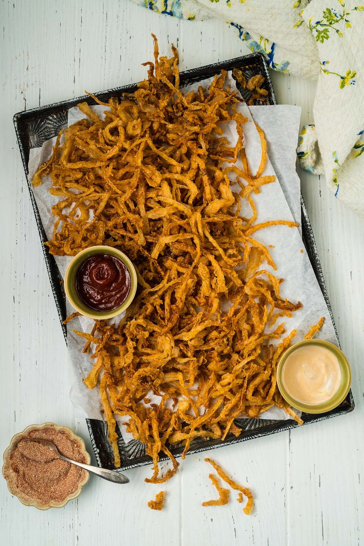 Crispy onion strings are piled high on a tray, served with ketchup, dipping sauce, and a small bowl of seasoning for the perfect snack.