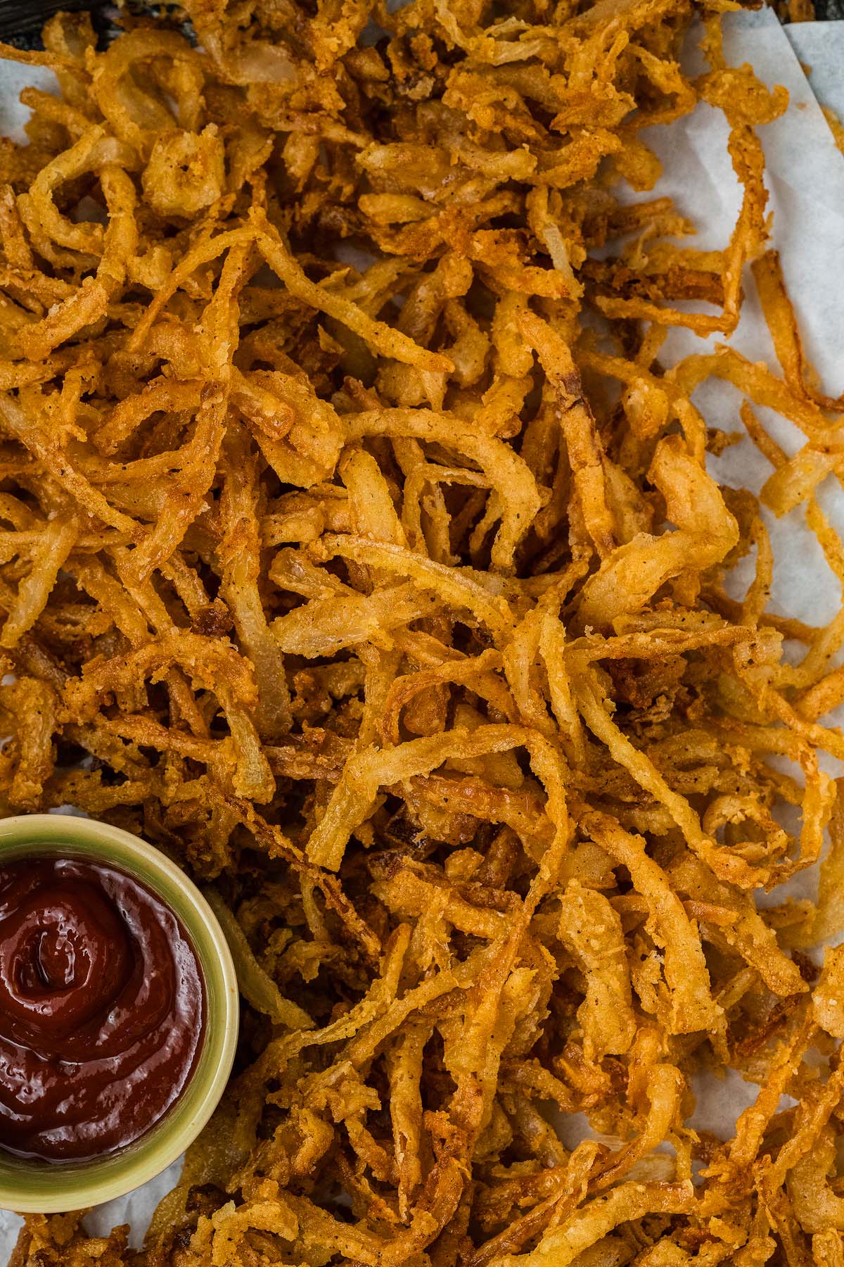 Crispy onion strings rest on parchment paper alongside a small bowl of ketchup for dipping.