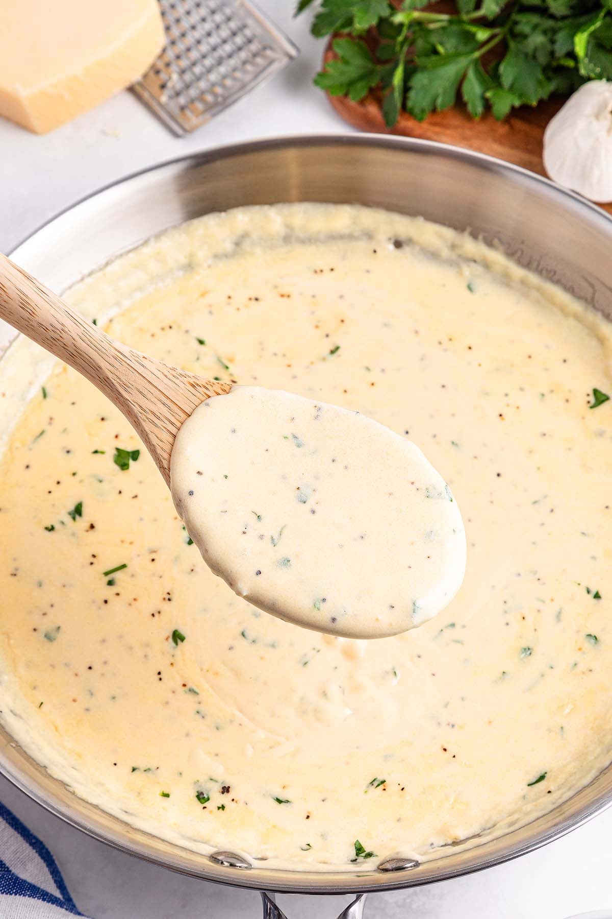 Wooden spoon scooping creamy white sauce with herbs from a pan, with parsley and cheese nearby.