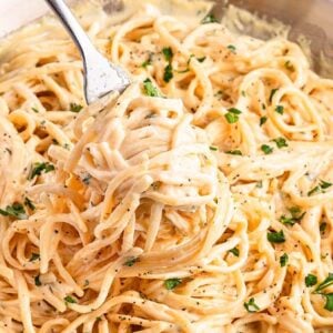 A fork twirls creamy fettuccine Alfredo garnished with chopped parsley in a pan.