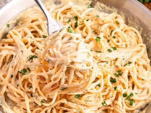 A fork twirls creamy fettuccine Alfredo garnished with chopped parsley in a pan.