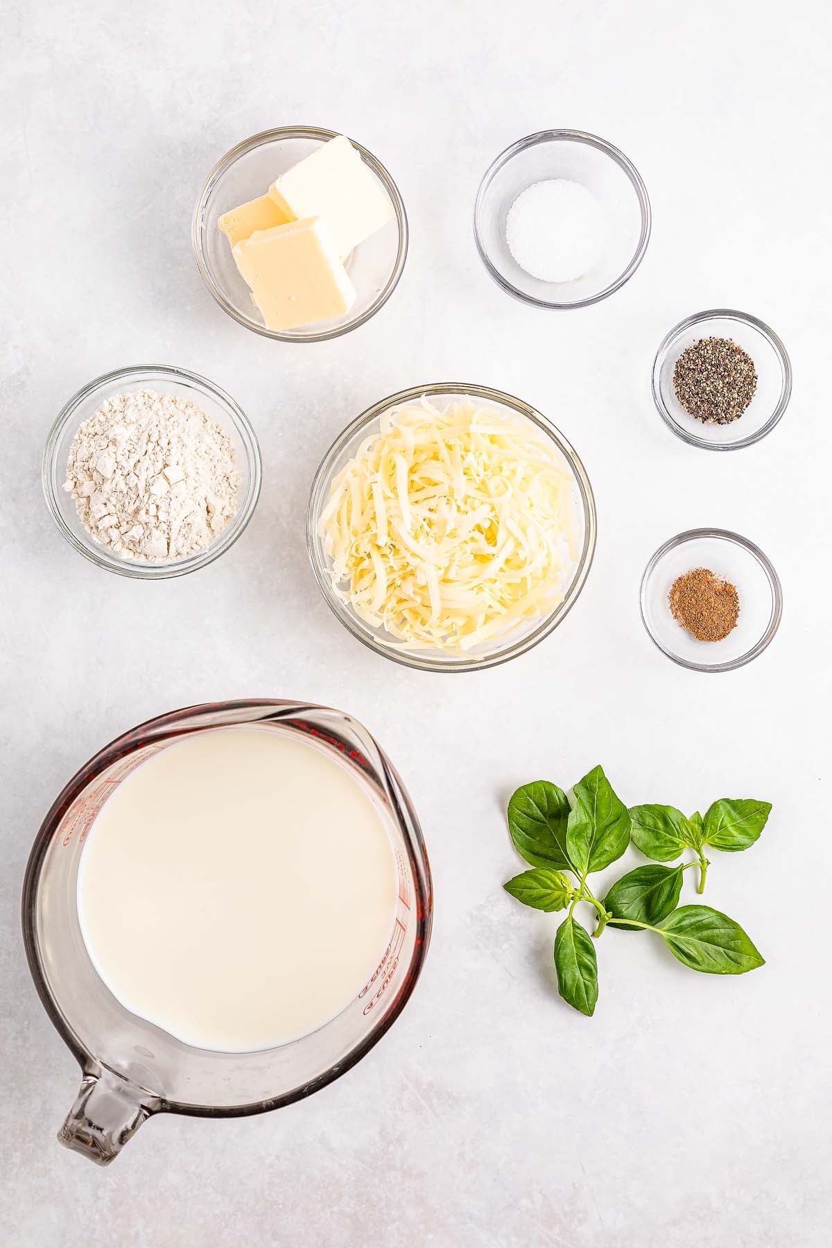 Ingredients for a sauce: milk, cheese, flour, butter, salt, pepper, nutmeg, and fresh basil on a white surface.