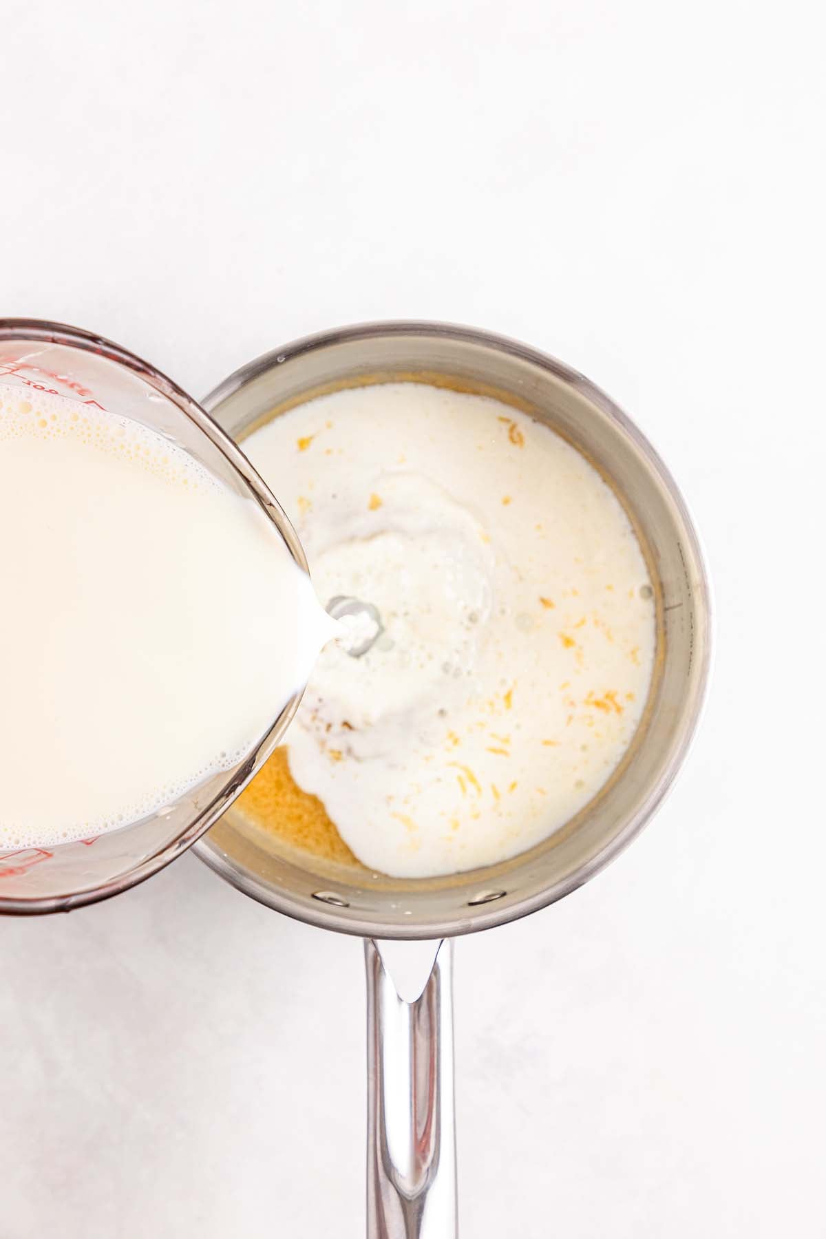 Milk is being poured from a measuring cup into a saucepan containing cheese and other ingredients.