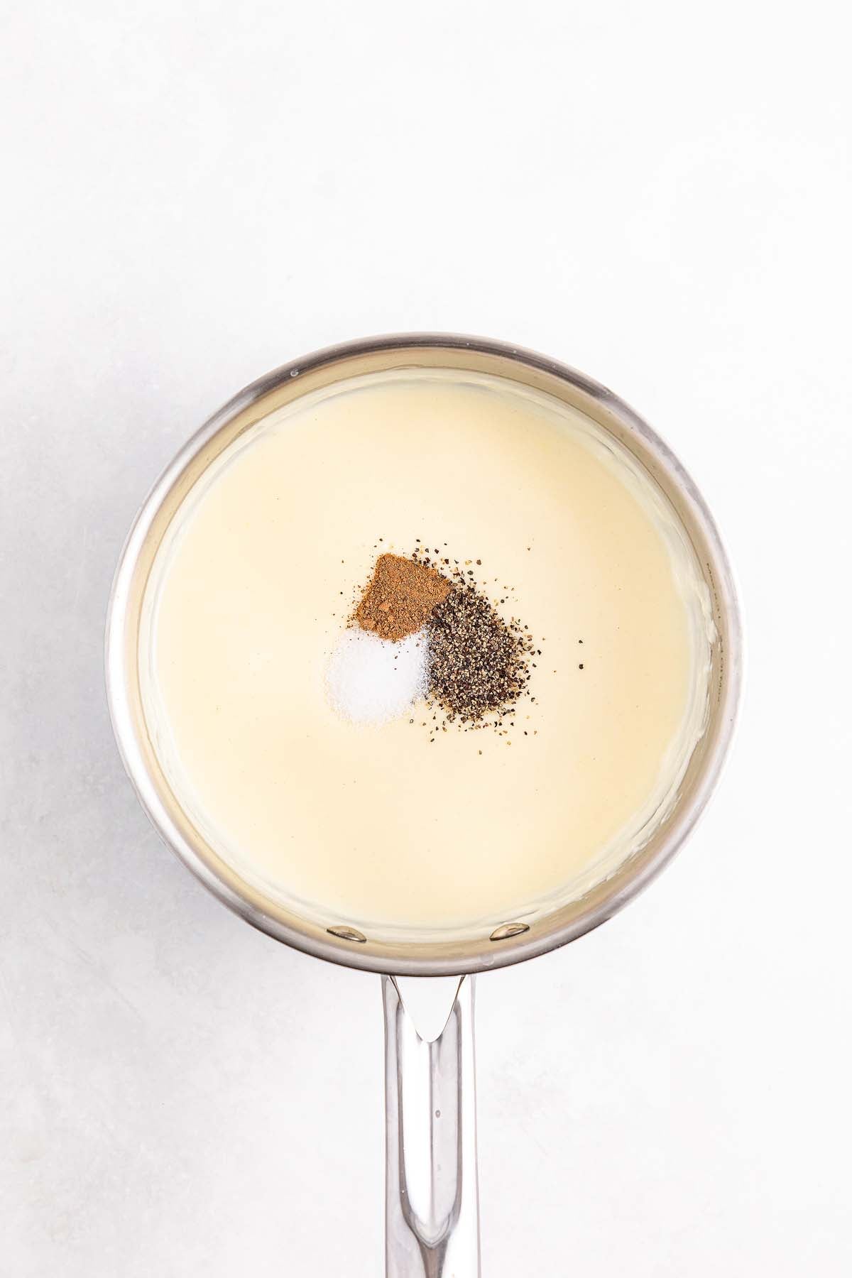 A saucepan filled with a creamy sauce, topped with salt, pepper, and spices on a white background.