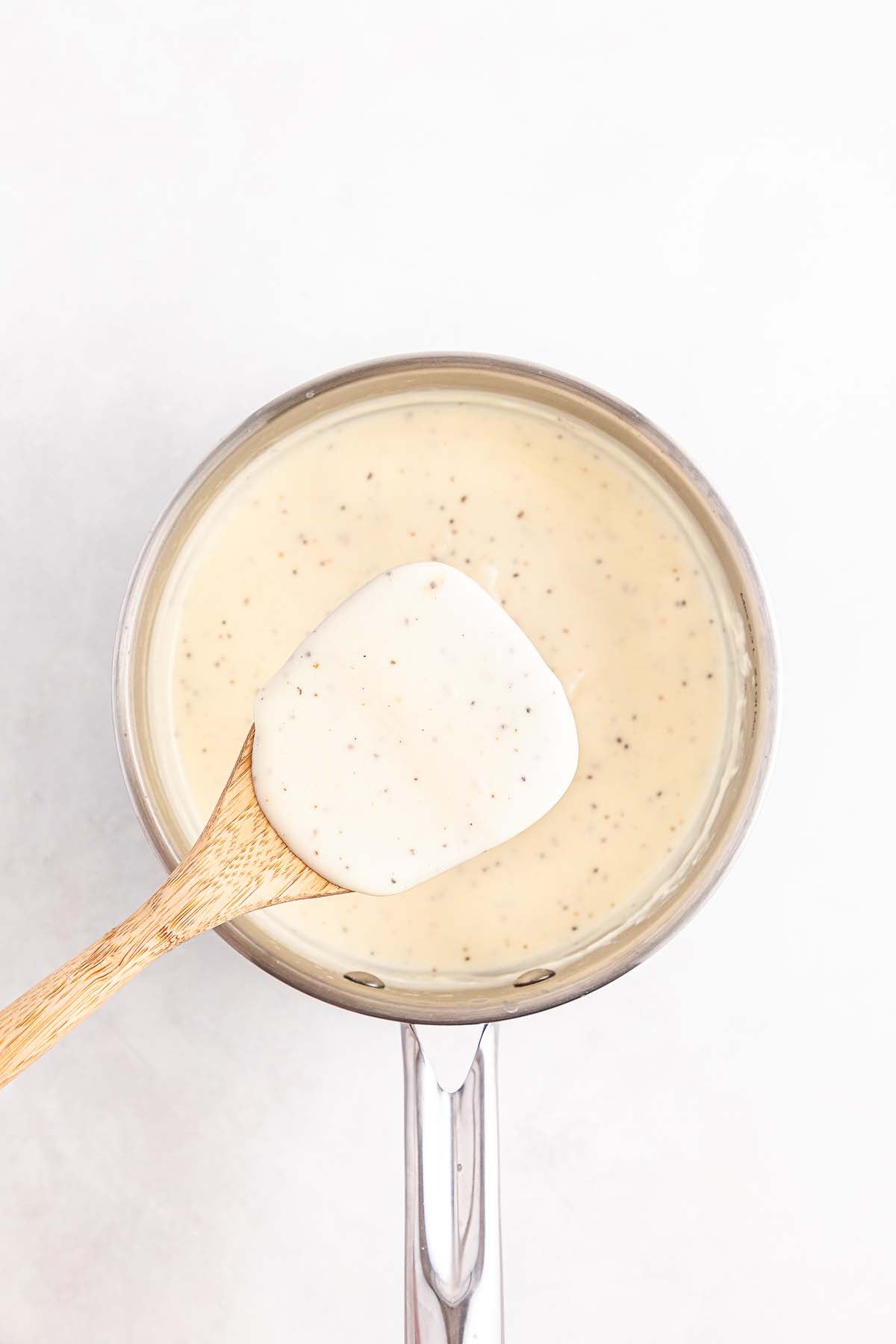 A saucepan of creamy white sauce with black pepper, stirred with a wooden spoon.