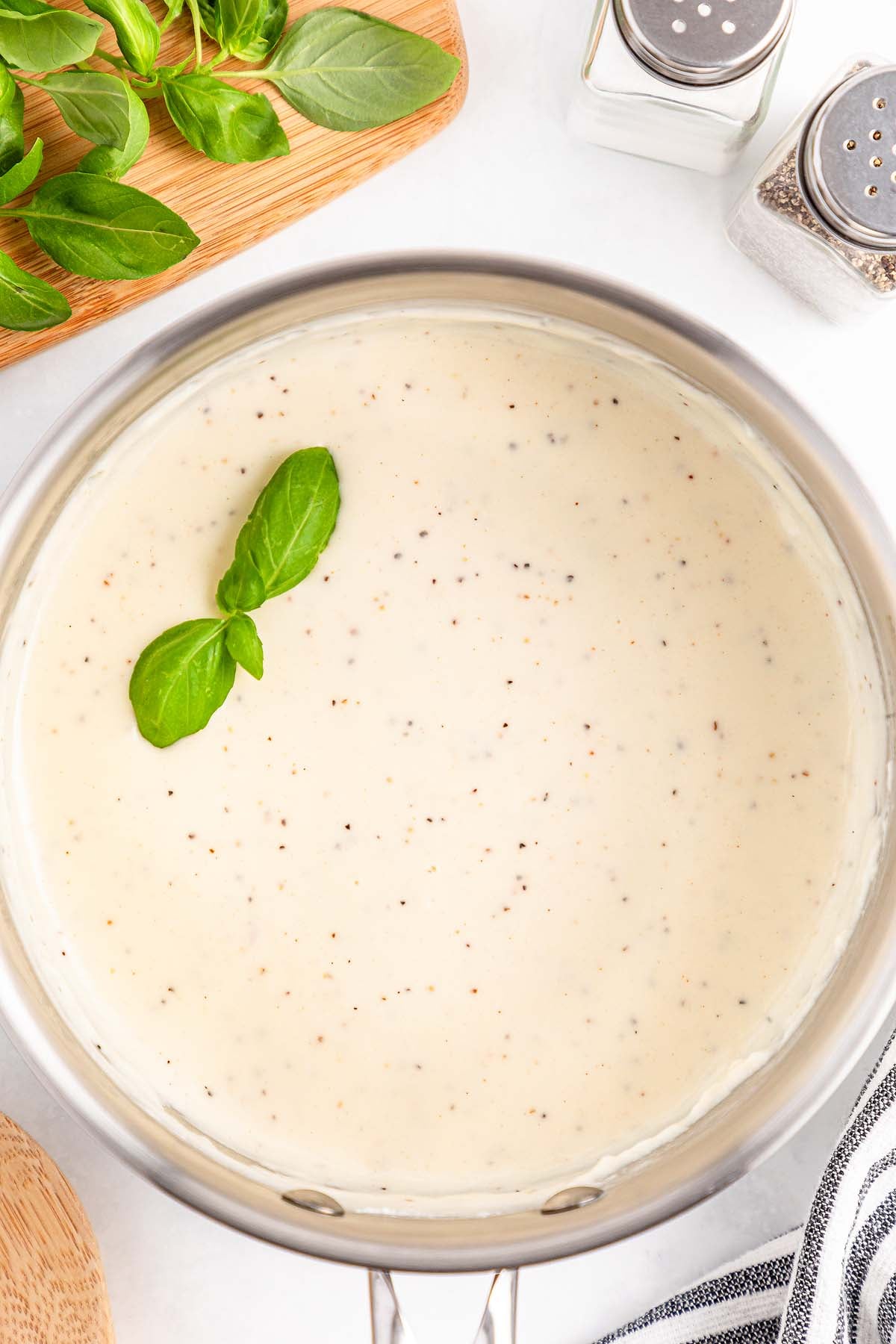 Creamy white sauce in a saucepan, topped with two basil leaves, with salt and pepper shakers nearby.