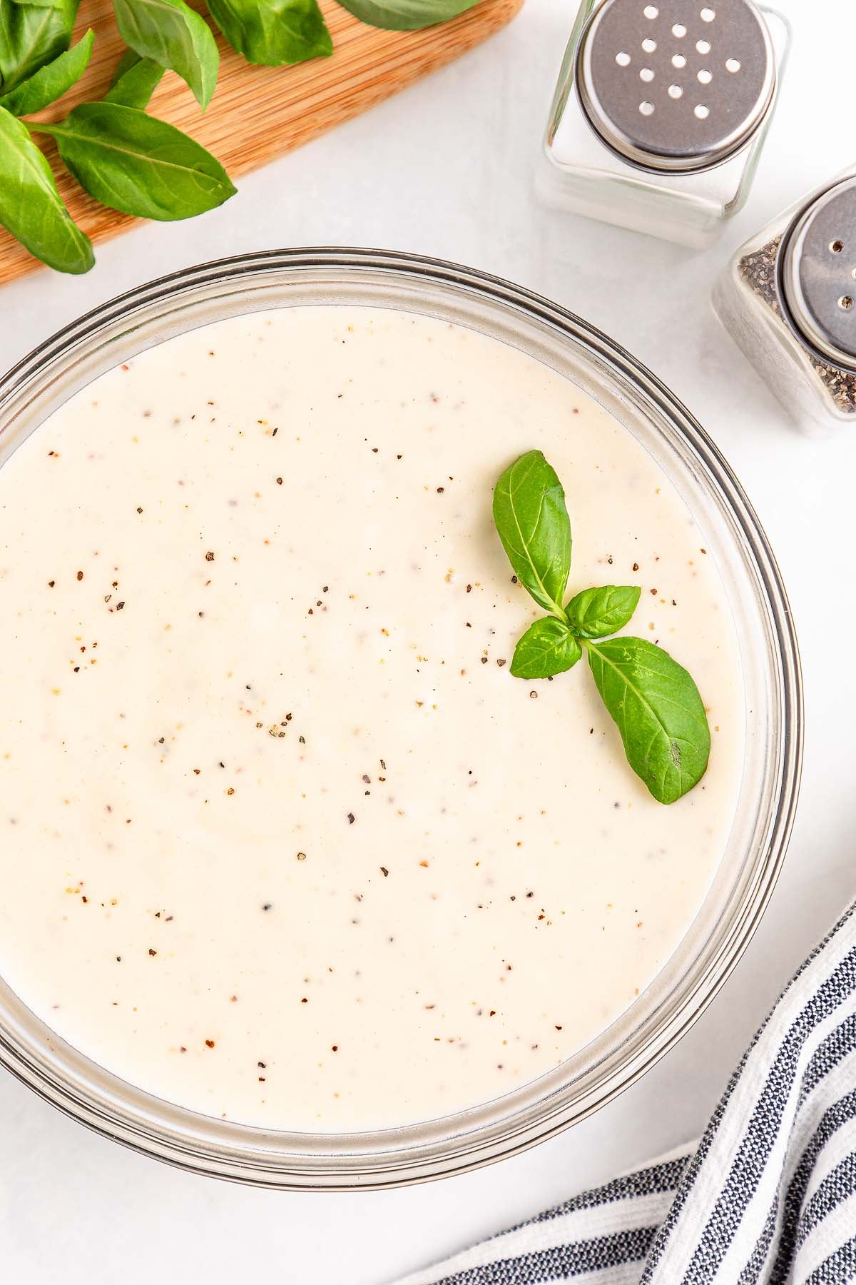 Creamy white sauce in a glass bowl, topped with basil leaves and black pepper, with seasonings nearby.