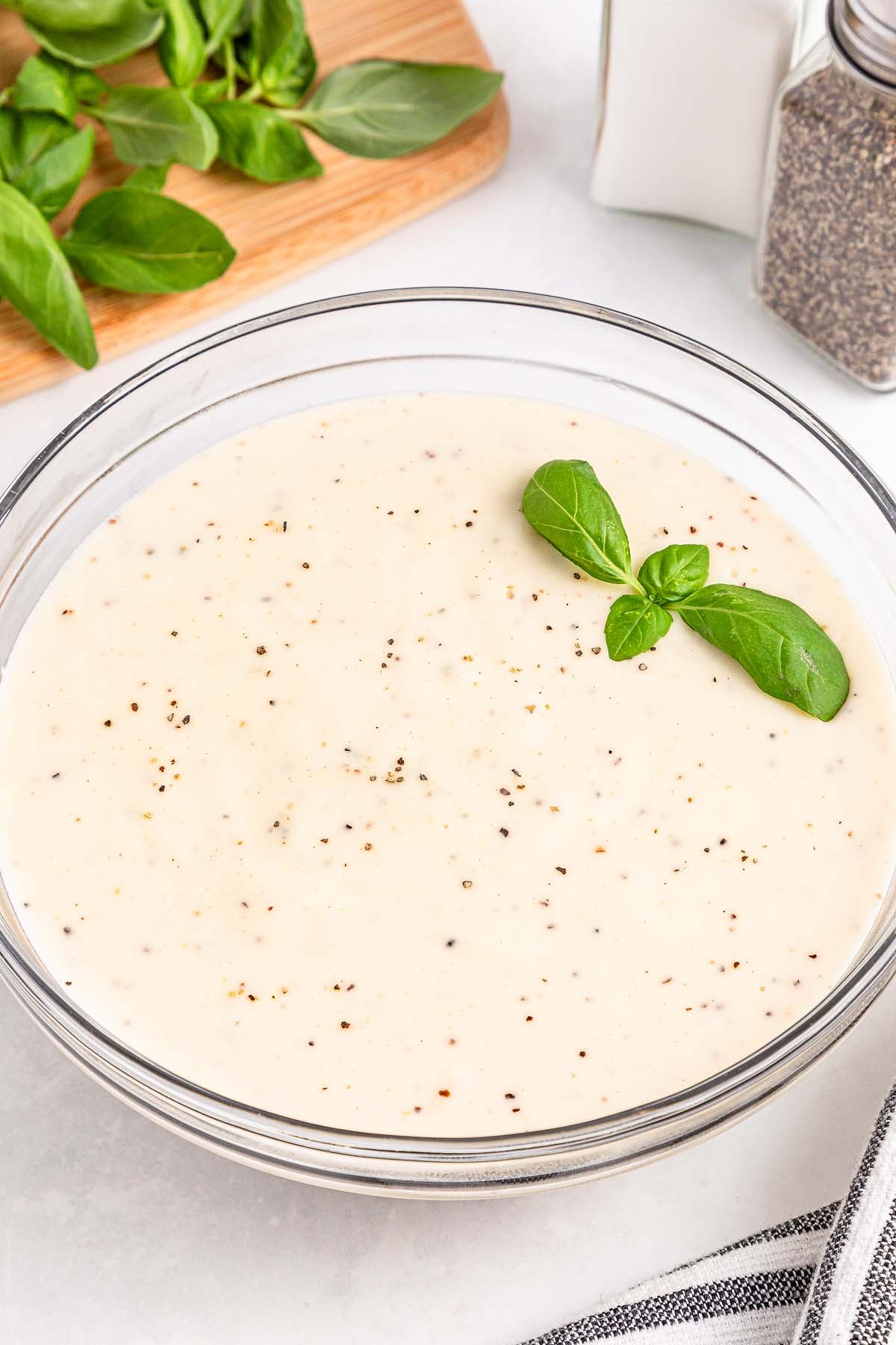 A bowl of creamy white sauce with black pepper and basil garnish, next to fresh basil and seasonings.