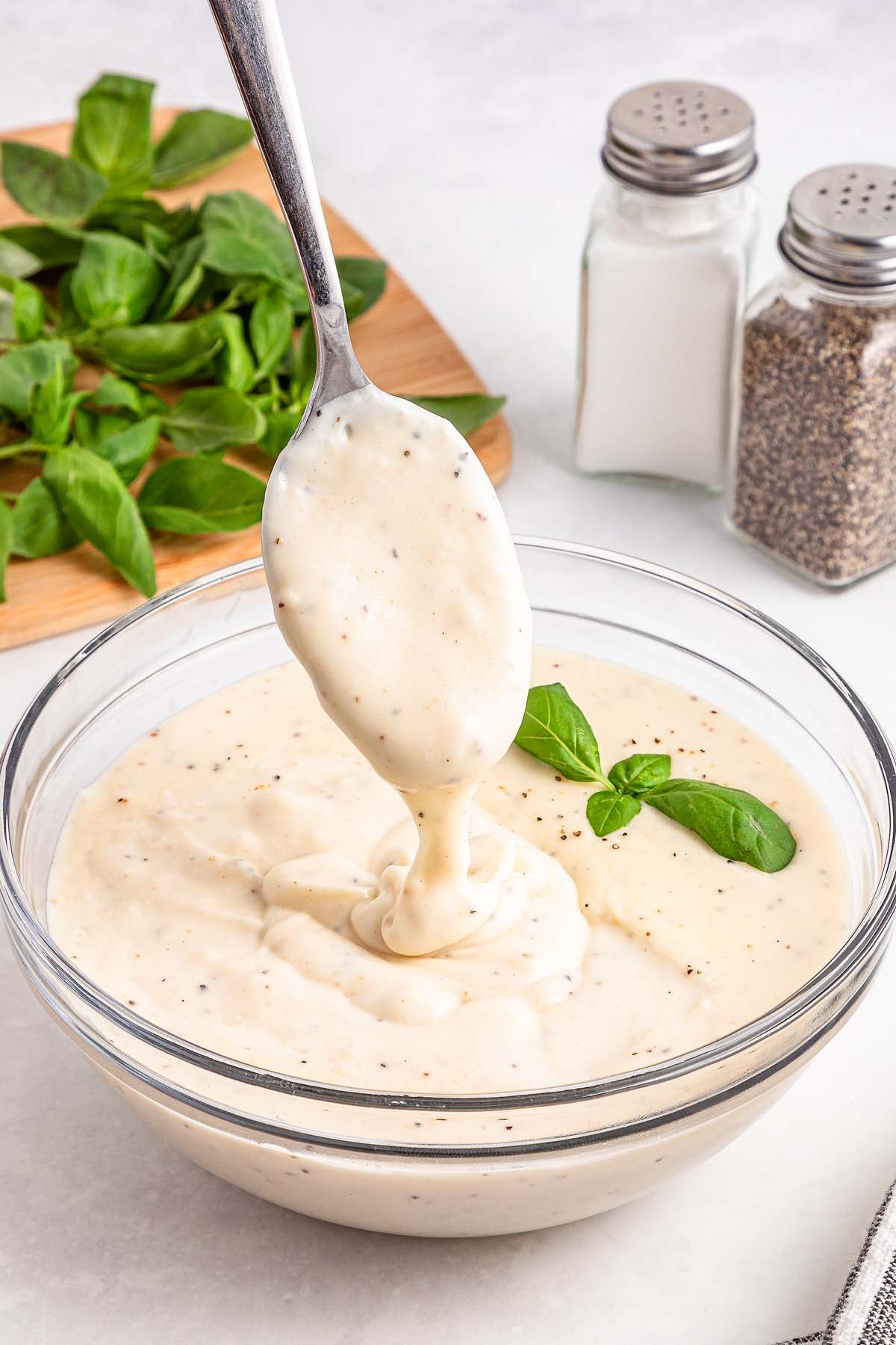 A spoon lifts creamy white sauce from a glass bowl, garnished with basil, with salt and pepper in the background.