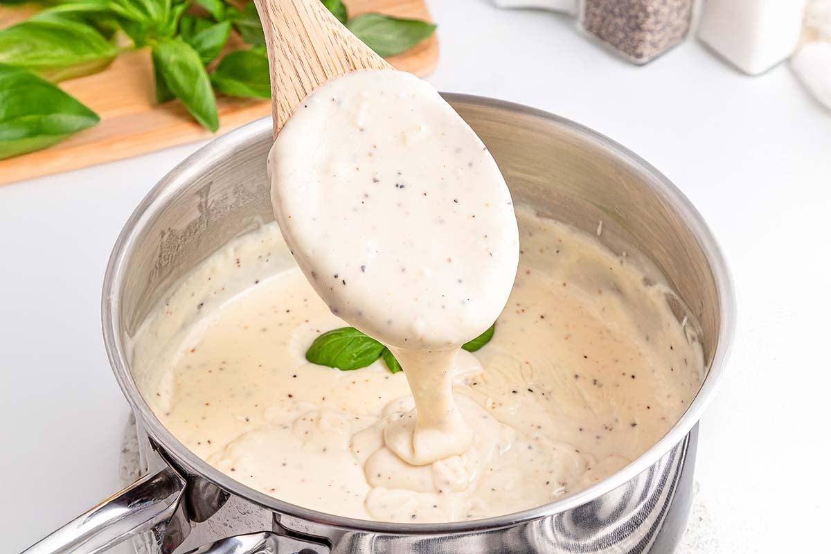 Wooden spoon lifts creamy white sauce with herbs from a stainless steel pot on a white kitchen counter.