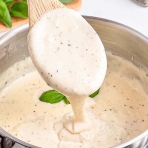 A wooden spoon lifts creamy white sauce with pepper from a saucepan, basil leaves in the background.