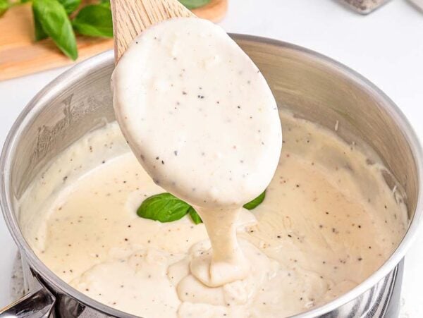 A wooden spoon lifts creamy white sauce with pepper from a saucepan, basil leaves in the background.