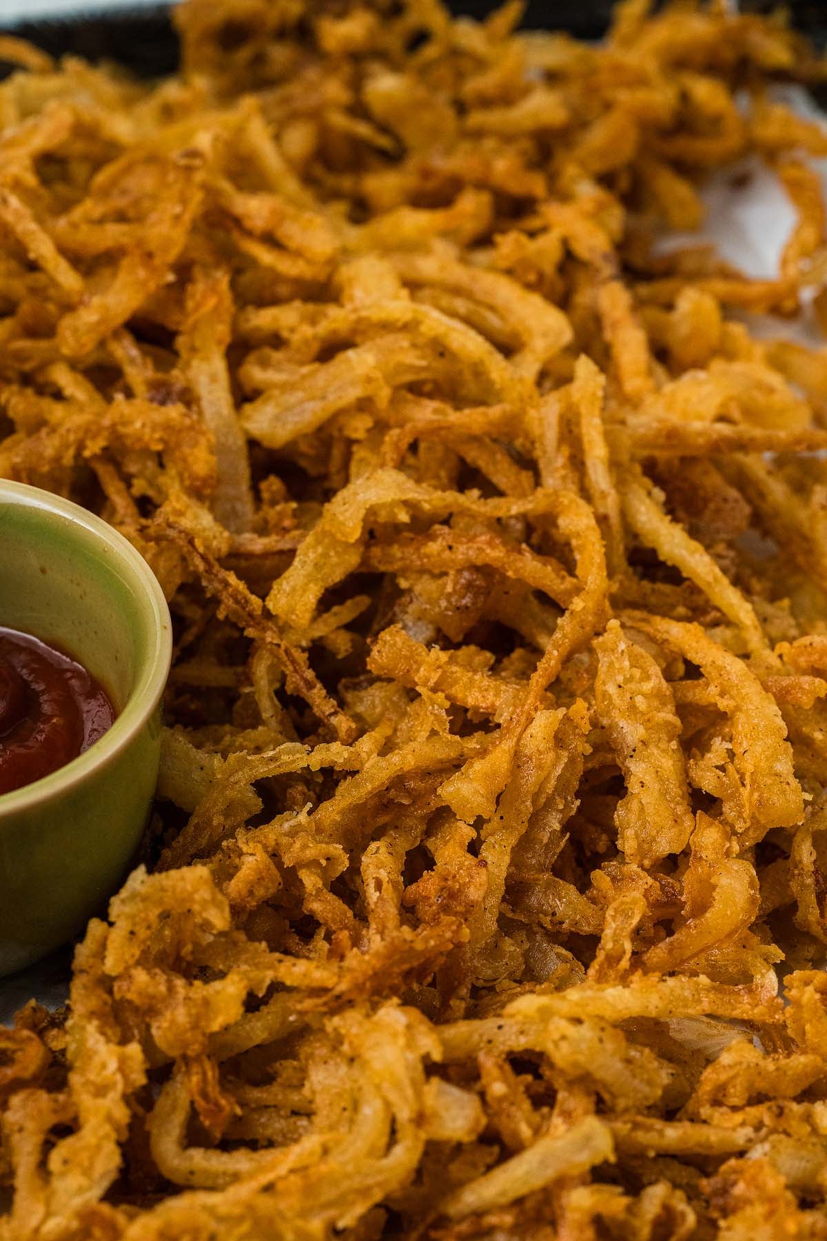 Crispy onion strings are piled high on a tray, served alongside a small bowl of ketchup for dipping.