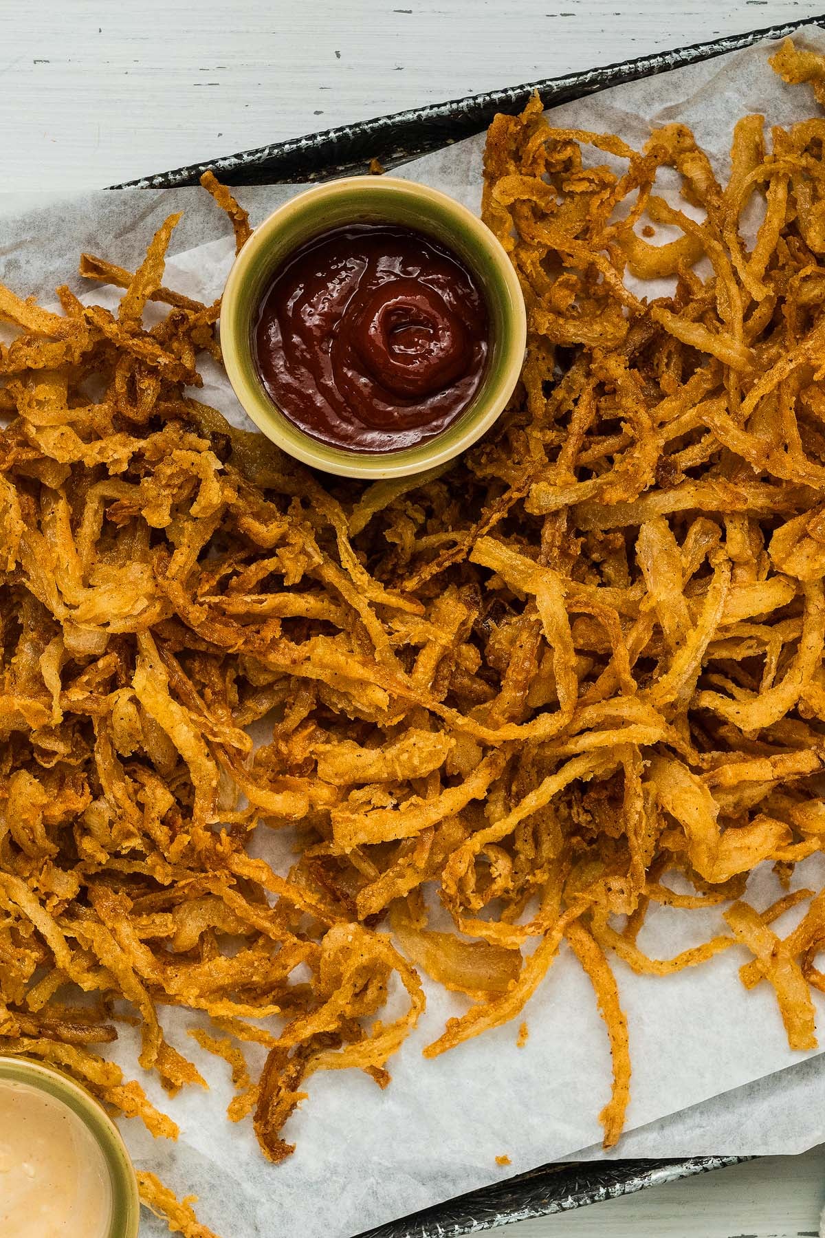 Crispy onion strings are piled high on parchment paper, served alongside a bowl of ketchup for dipping.