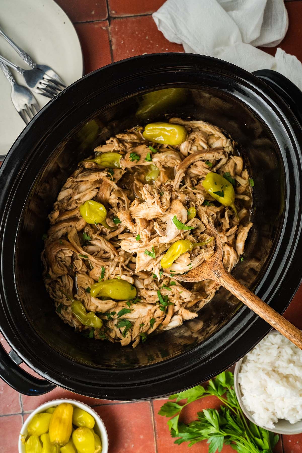 Shredded chicken with peppers in a slow cooker, served with rice, parsley, and utensils on a red tiled table.
