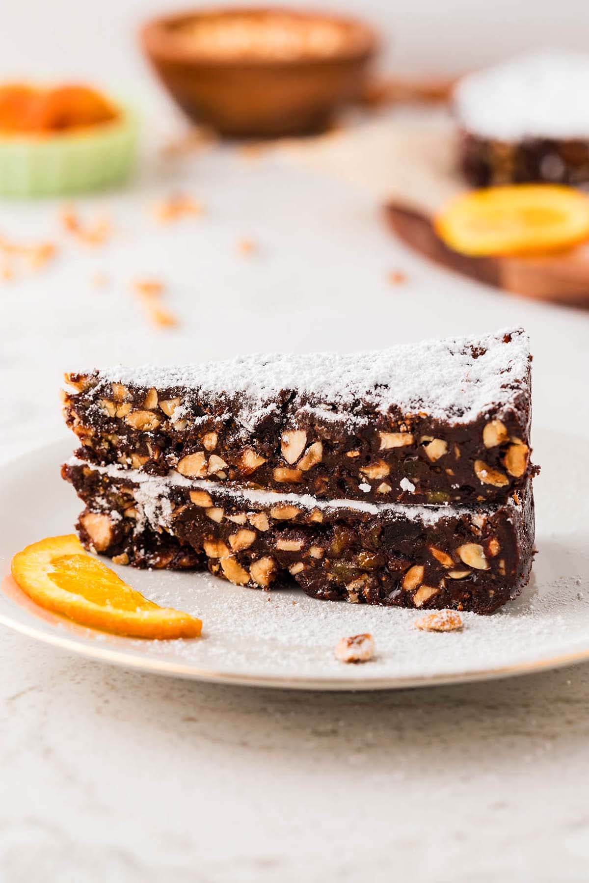Two slices of chocolate nut cake dusted with powdered sugar, garnished with an orange slice on a plate.